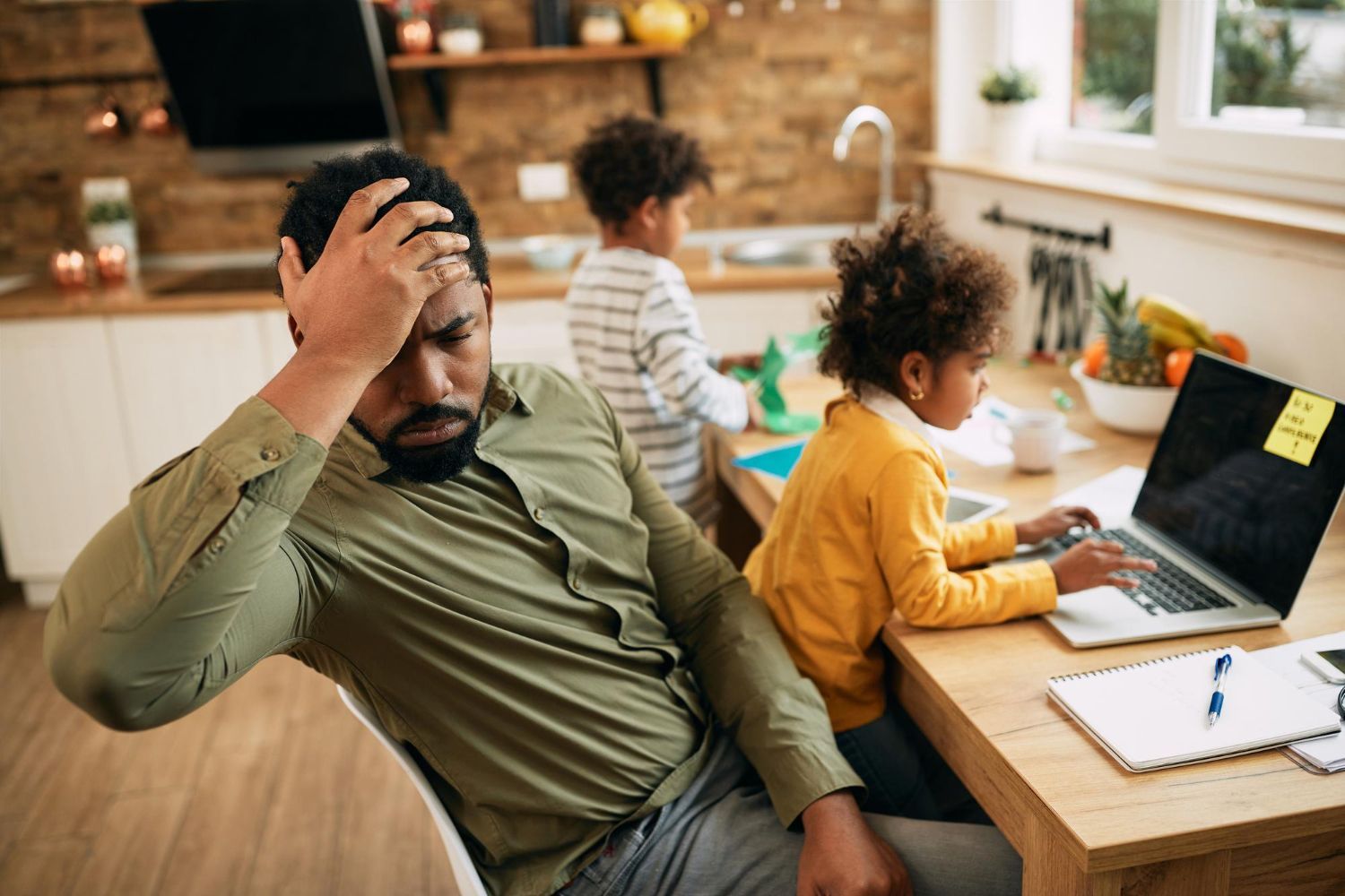 Man at kitchen table, stressed, hand on head, children in background, working at laptop.