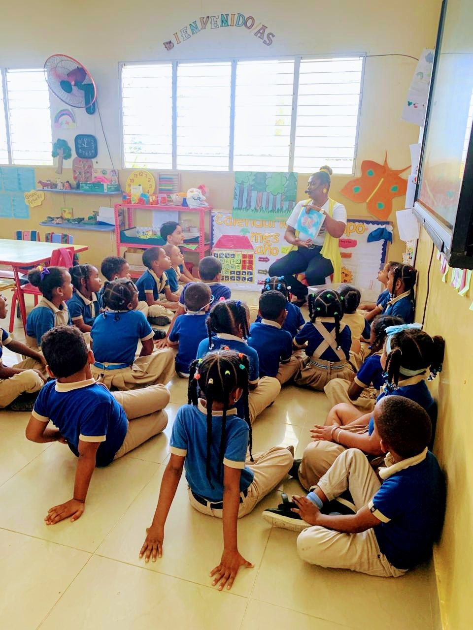 Children in blue shirts listen to a teacher read a book in a brightly colored classroom.