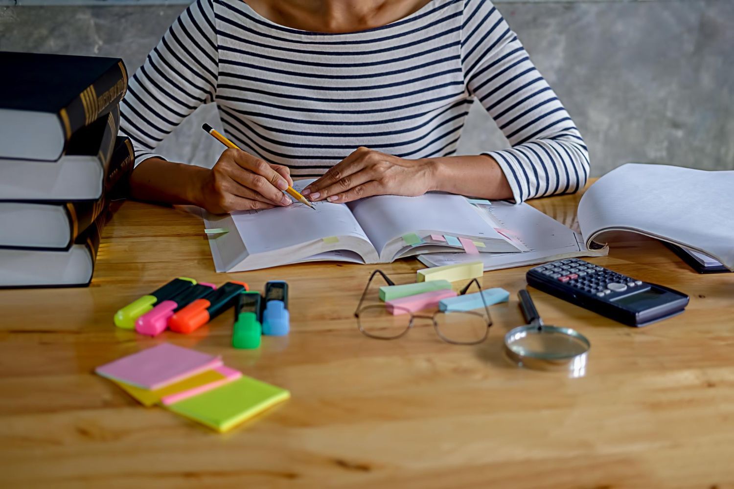 Woman in striped shirt writing in open book at a wooden desk with books, highlighters, calculator.