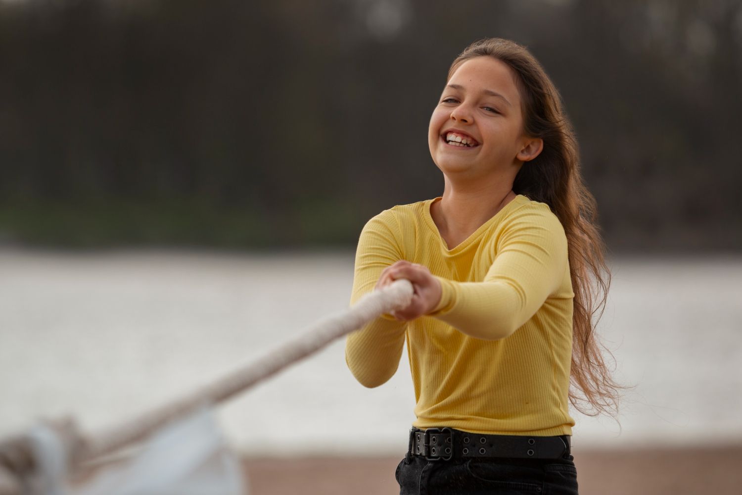 Girl in yellow shirt laughs, pulling a rope outdoors near water.