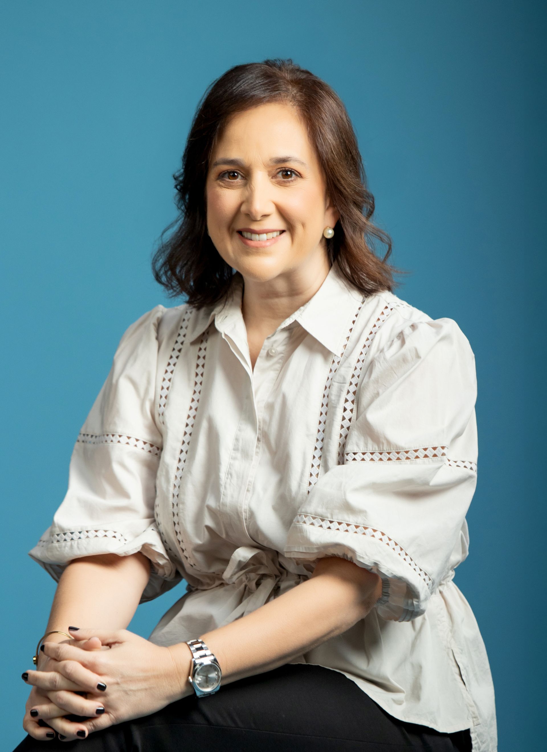 Woman with dark hair, smiling, wearing a white blouse, blue background.