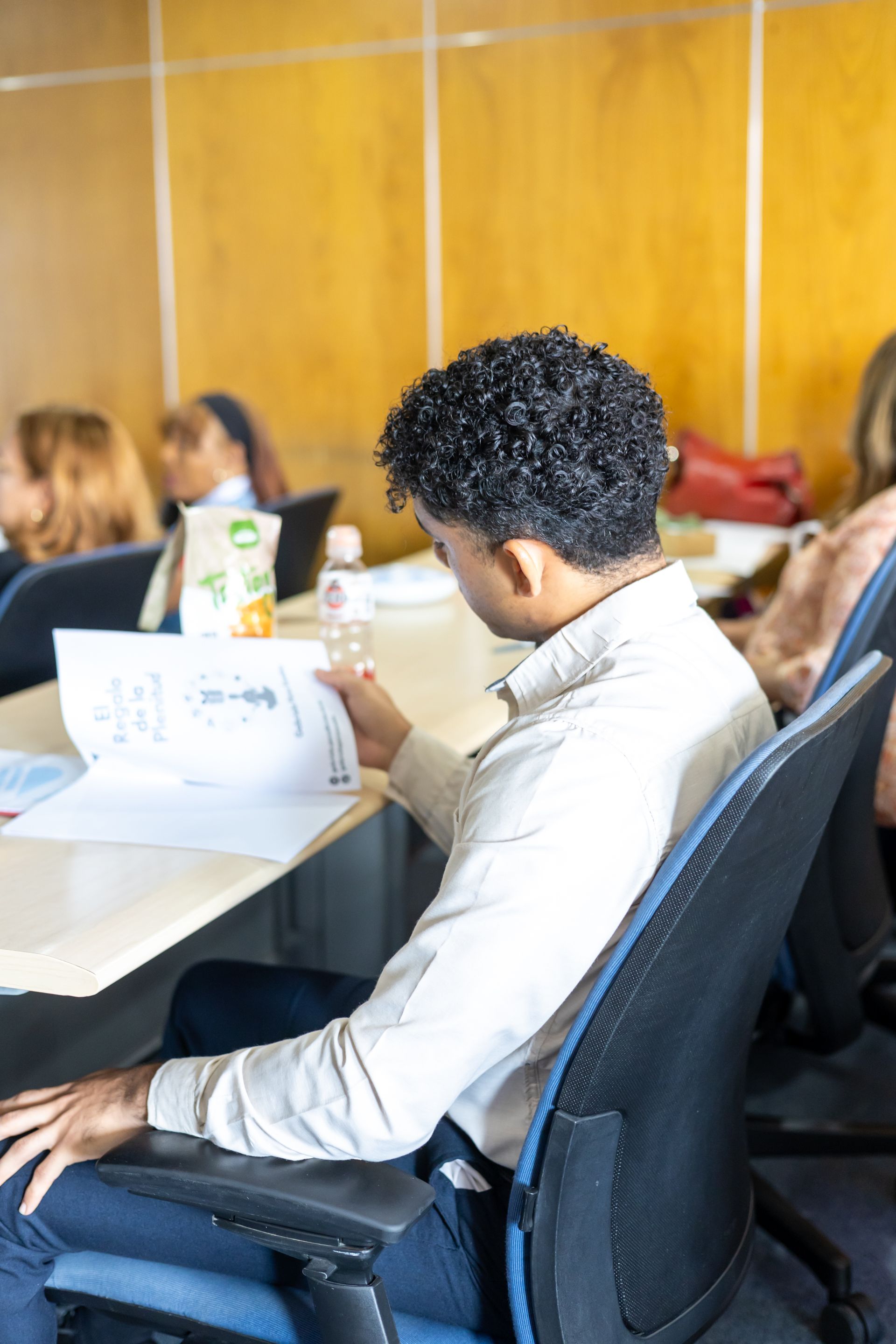 Person in a light-colored shirt studying papers at a table in a classroom setting. Others sit nearby.