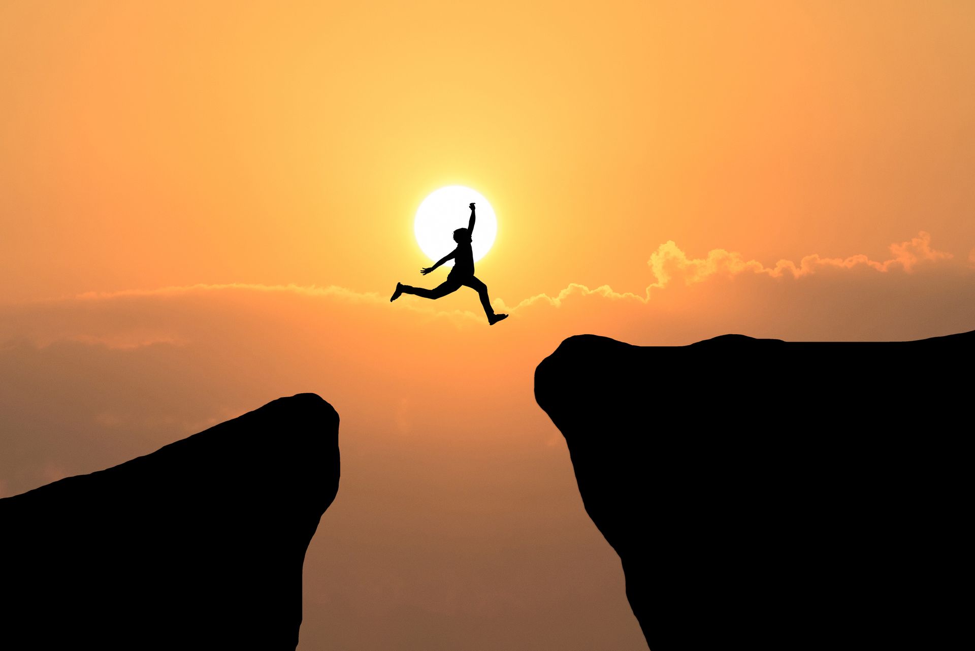 Silhouette of a person leaping over a chasm at sunset, arms raised in triumph.