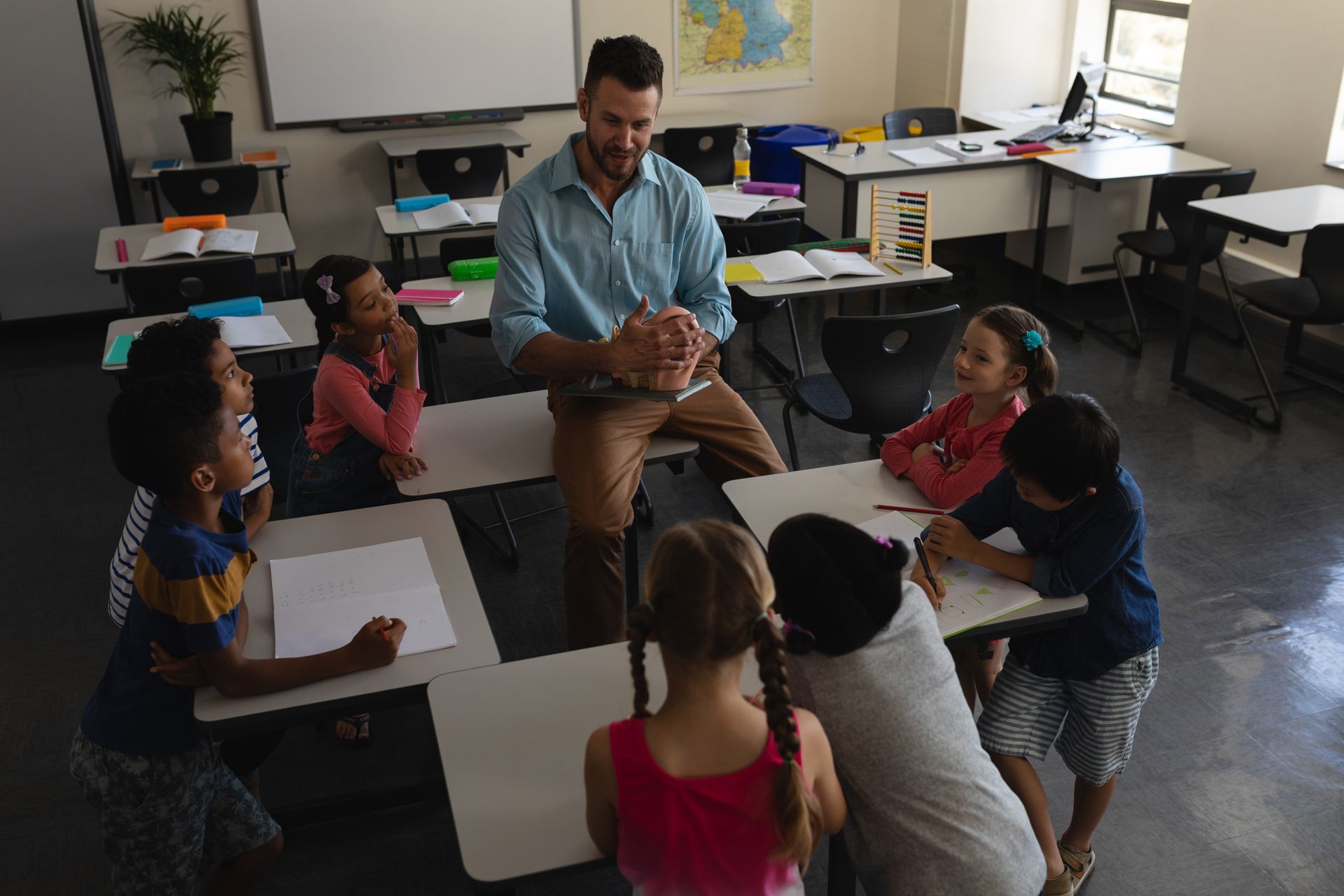 Teacher in classroom with children, sitting around desks, smiling.