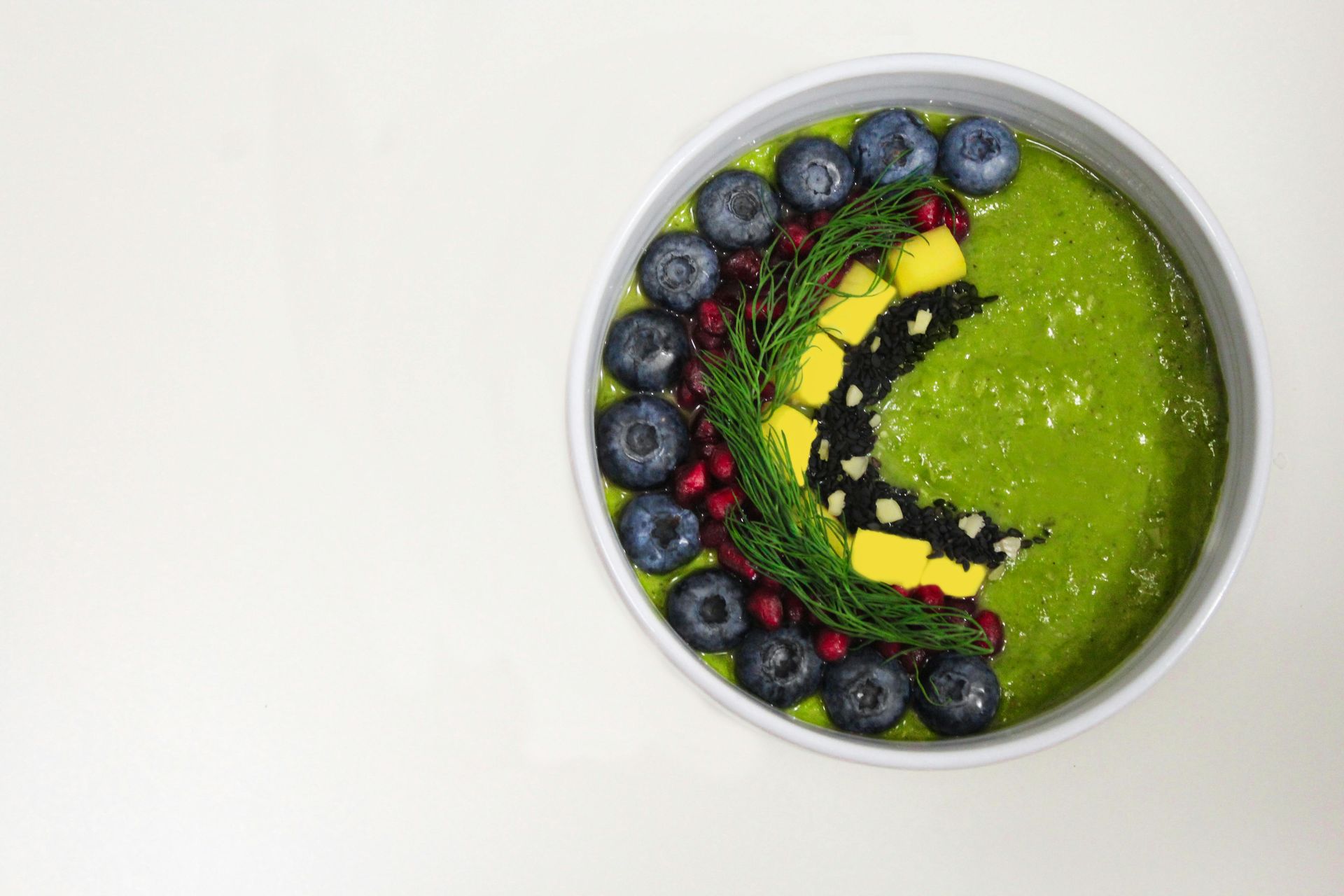 A bowl of green smoothie with blueberries , mango , and dill on a white surface.