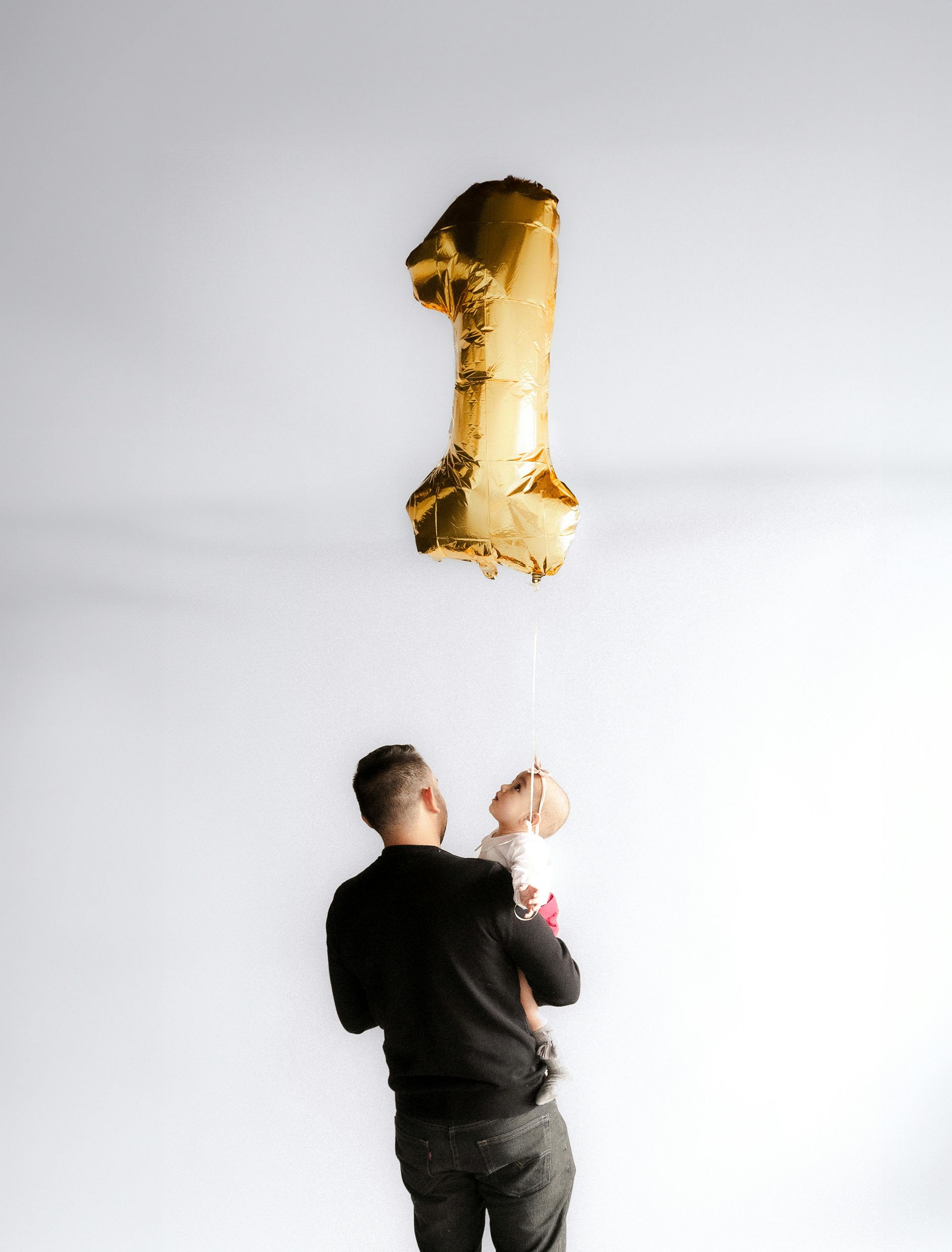 A man is holding a baby and a balloon in the shape of the number 1