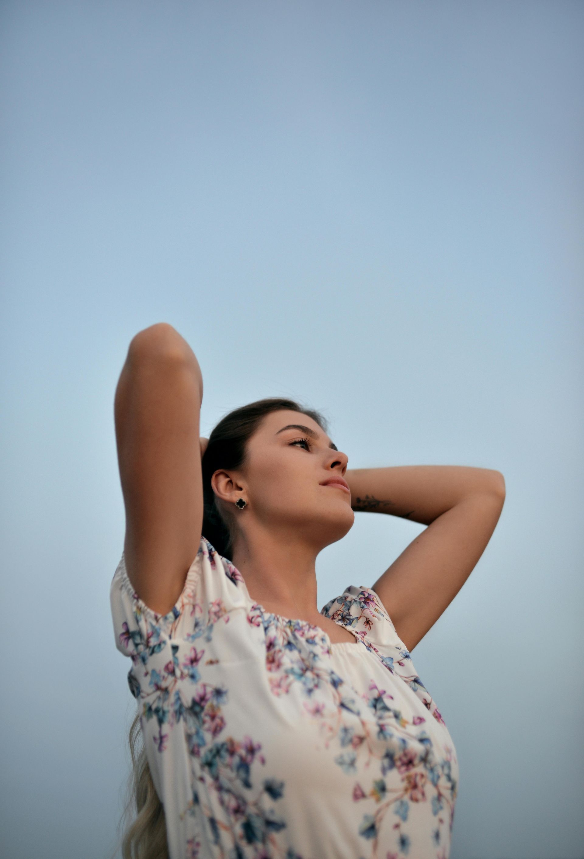 Woman with hands behind her head, looking up. She wears a floral dress, against a blue sky background.