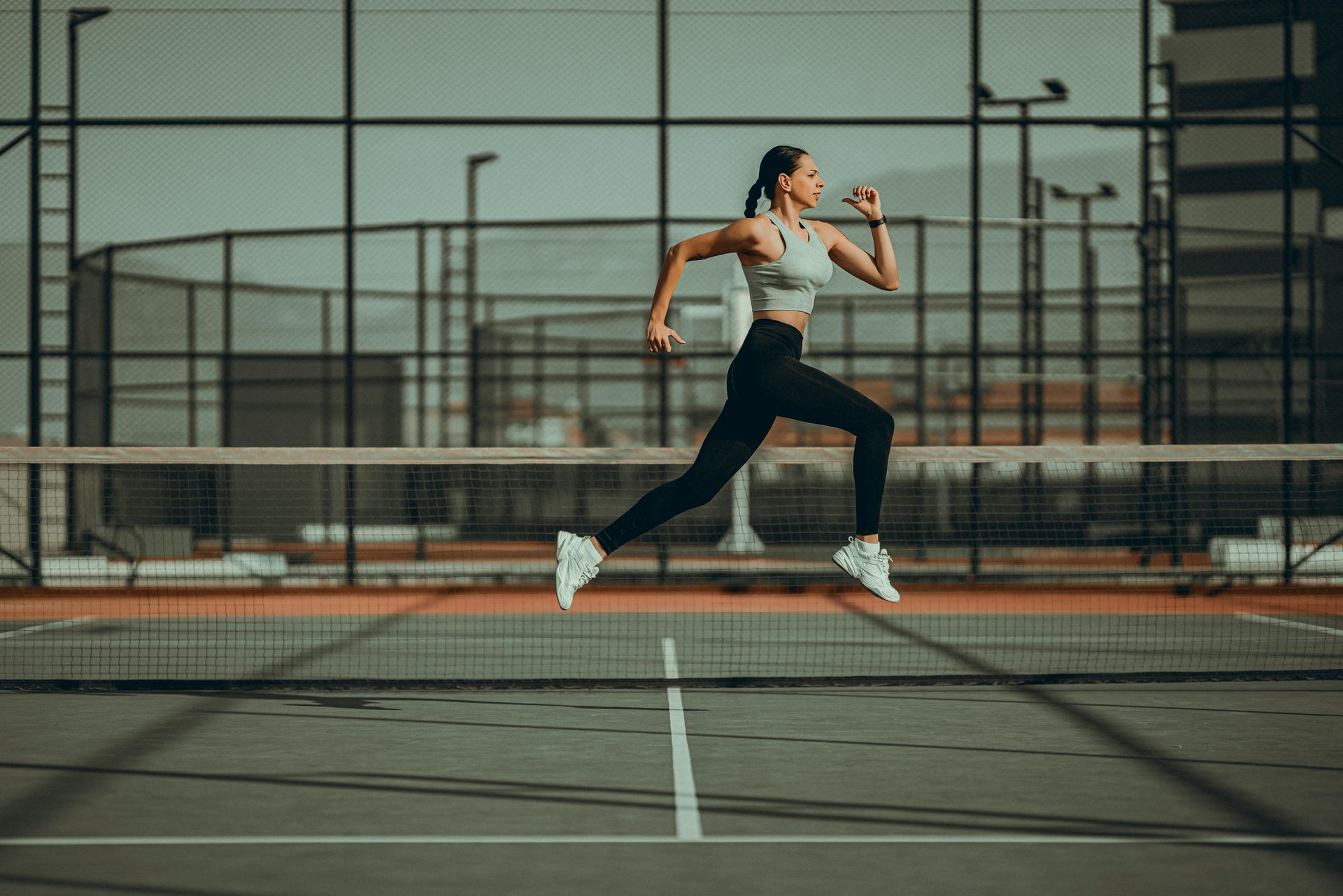 Woman running on a tennis court wearing athletic clothing; city backdrop.