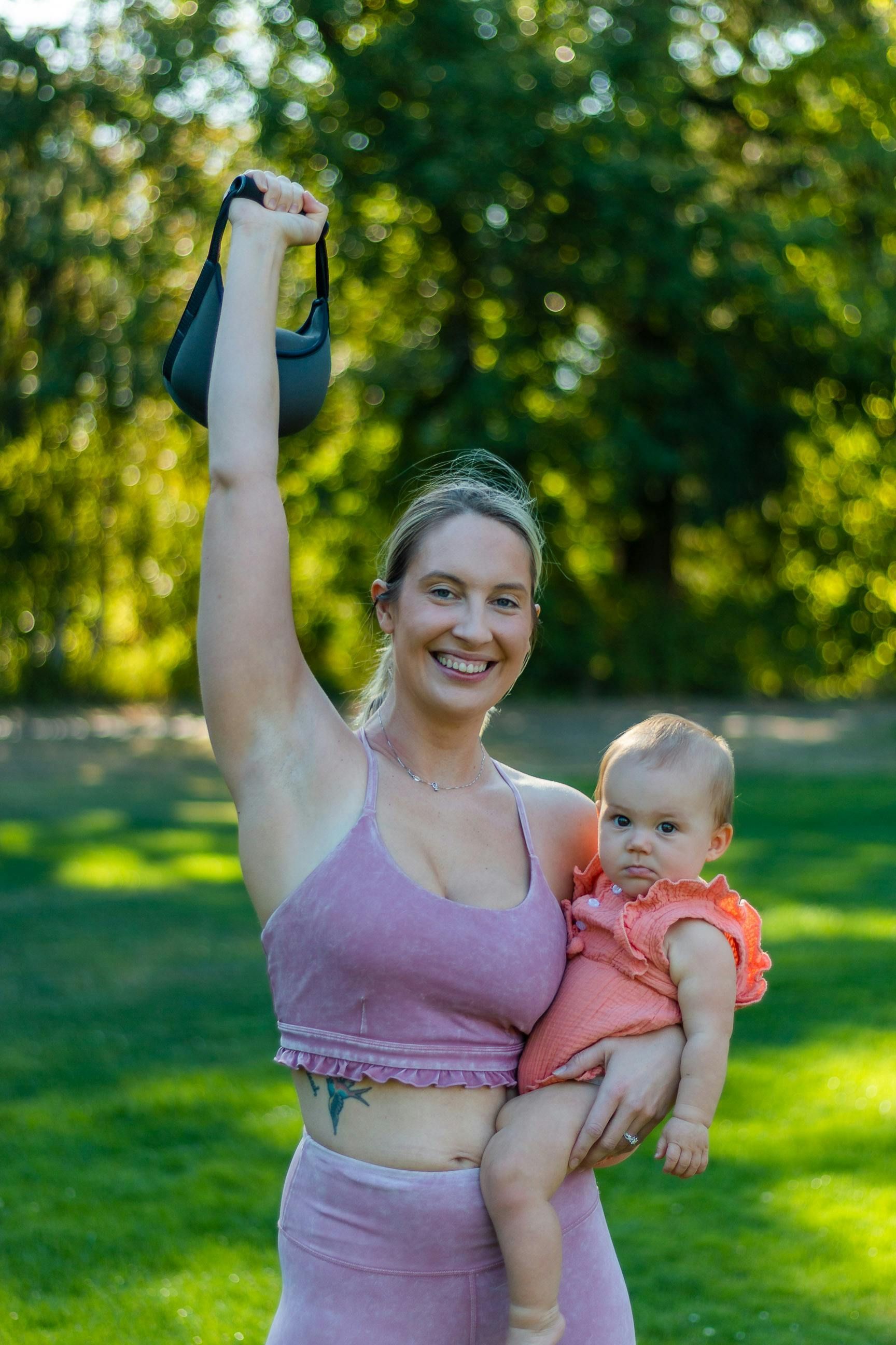 A woman is holding a baby and a kettlebell in a park.