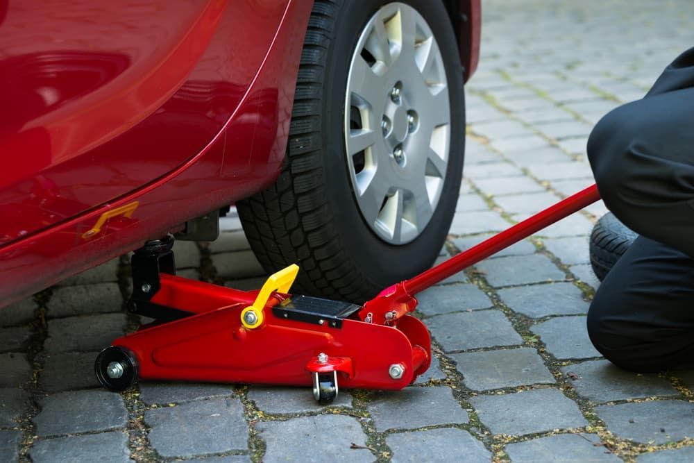 Red Car Being Lifted by a Red Floor Jack on a Cobblestone Surface; a Person Kneels Nearby — FNQ Filters & Parts In Mareeba, QLD