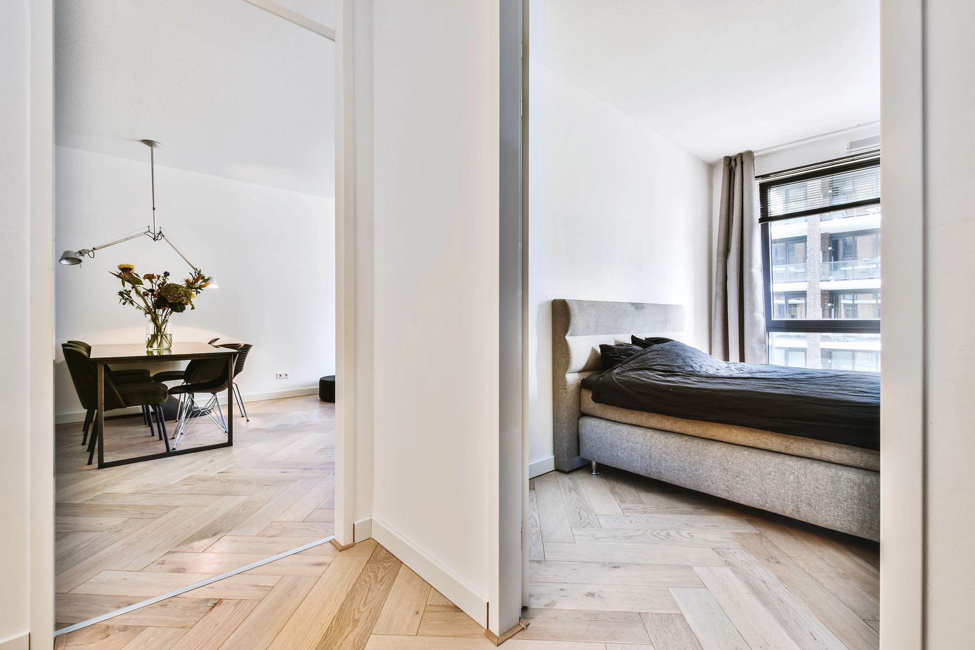 La porta aperta rivela la zona pranzo con tavolo e la camera da letto con letto. Pavimento in legno chiaro.