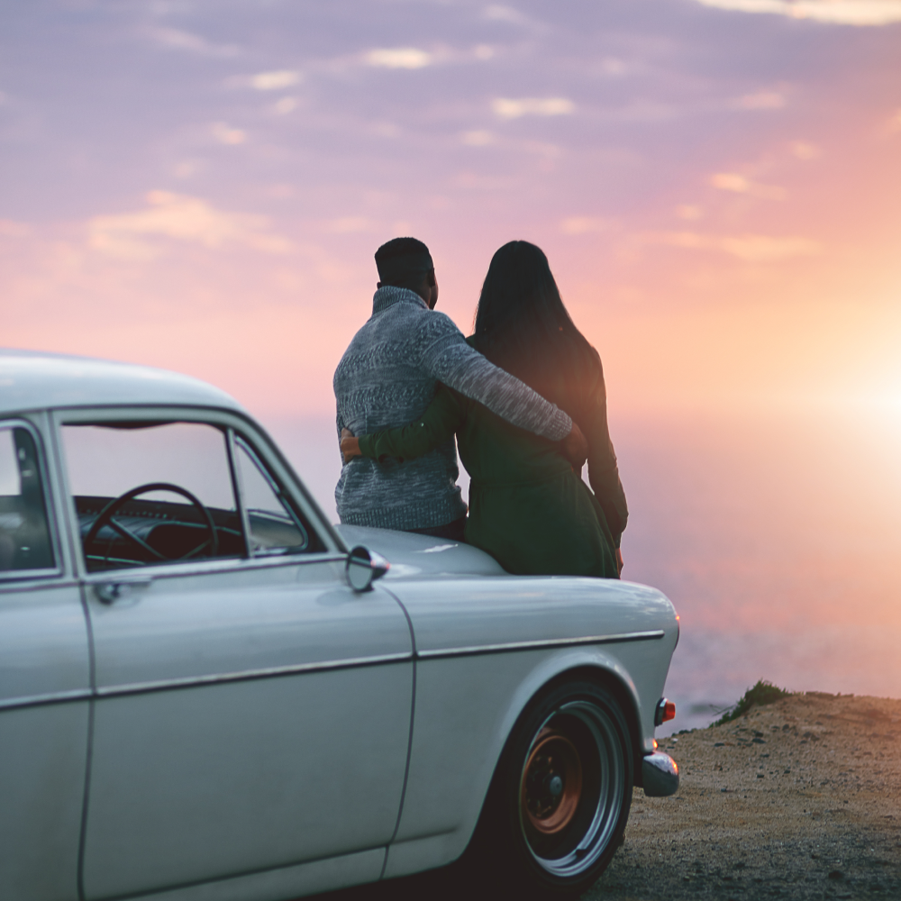 A man and woman are sitting on the back of a car looking at the sunset