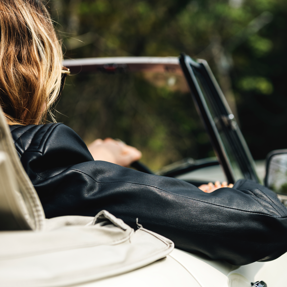 A woman in a black leather jacket is driving a white car