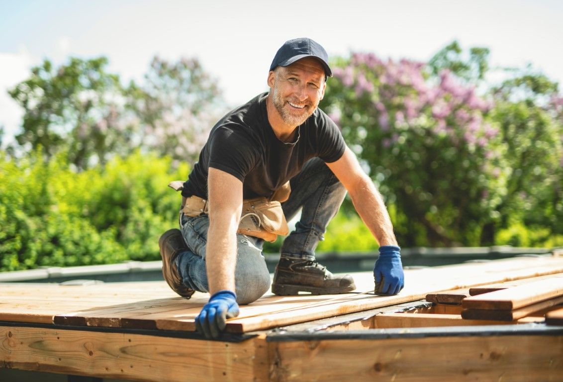 Builder kneeling on a wooden deck frame outdoors, wearing a black shirt, cap, and blue gloves.
