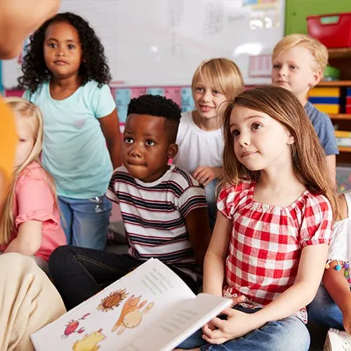 A group of children are sitting on the floor listening to a teacher read a book.