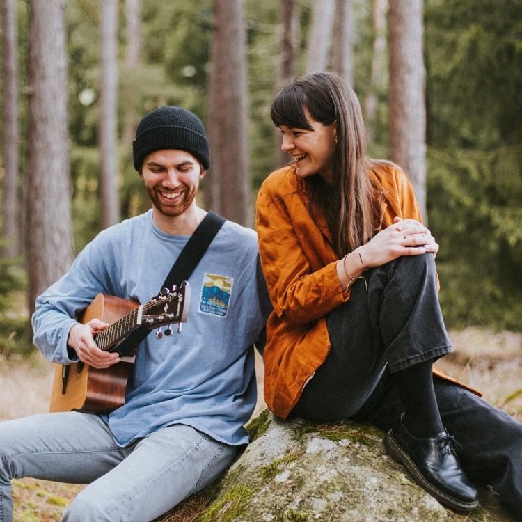 die beiden Musiker von der Indie-Band Ines mag's sitzen auf einem Stein im Wald. Markus hat seine Gitarre in der Hand, ein blaues T-Shirt an und eine schwarze Haube auf, während Karin ihn mit langen dunklen Haaren, einem orangenen Hemd und schwarzer Hose ansieht.