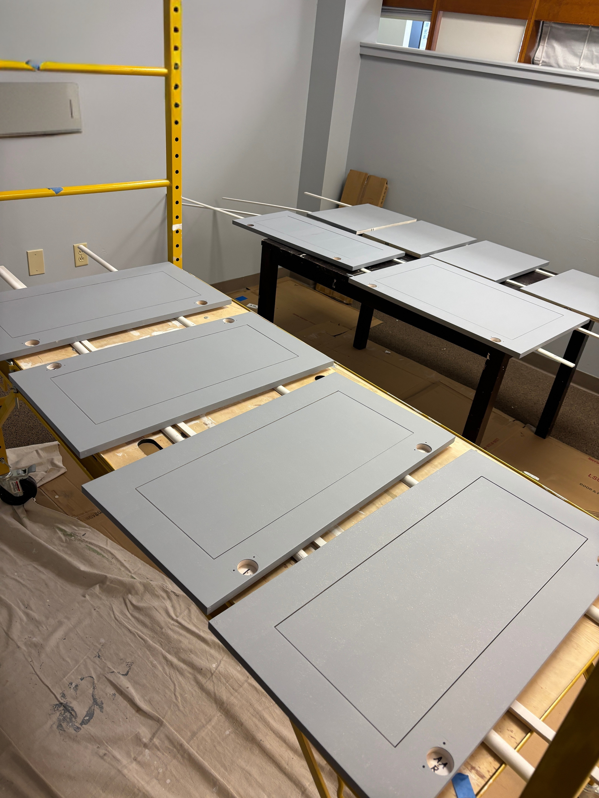 Gray cabinet doors and panels laid out on tables in a workshop, with a yellow ladder and shelves nearby.