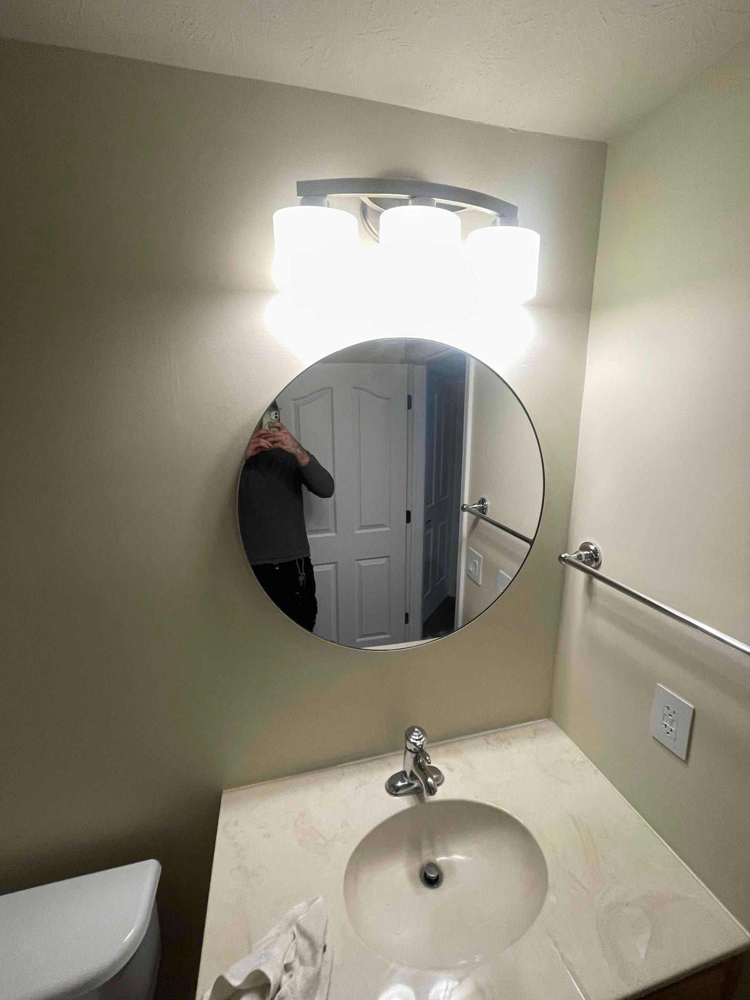Small bathroom vanity with round mirror, bright lights, sink, and towel bar against beige walls.