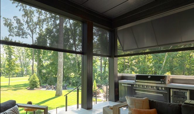 Covered patio with grill, lounge seating, and large windows overlooking a green yard and trees.