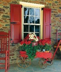 Red flower-filled wagon and chair on a porch beneath a window with red shutters