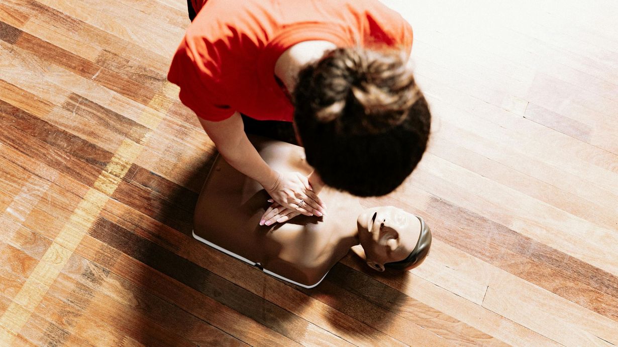 Person in an orange shirt giving CPR to a manikin on a wooden floor.