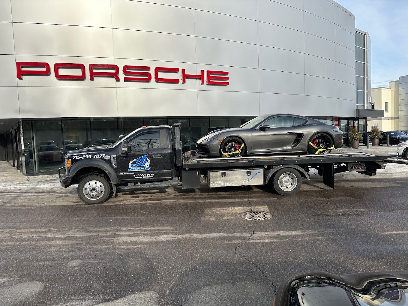 A tow truck is towing a car in front of a porsche dealership.