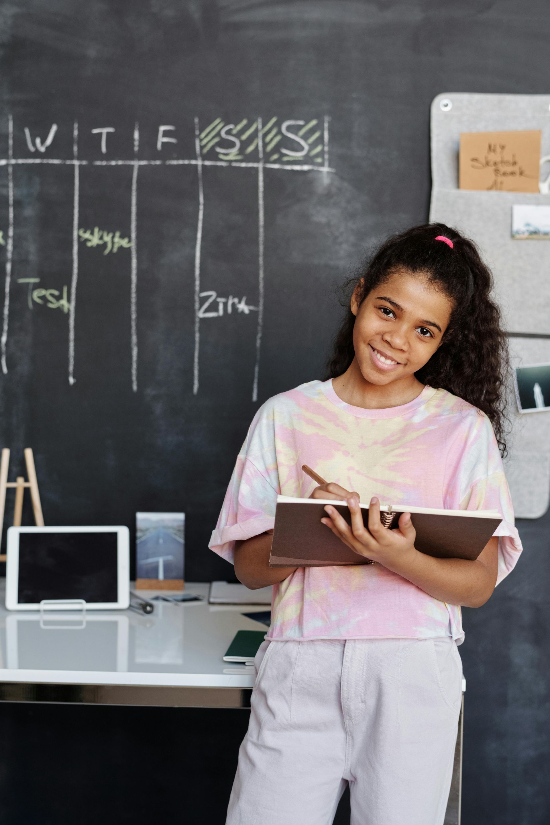 A young girl is standing in front of a blackboard holding a notebook and a pen.