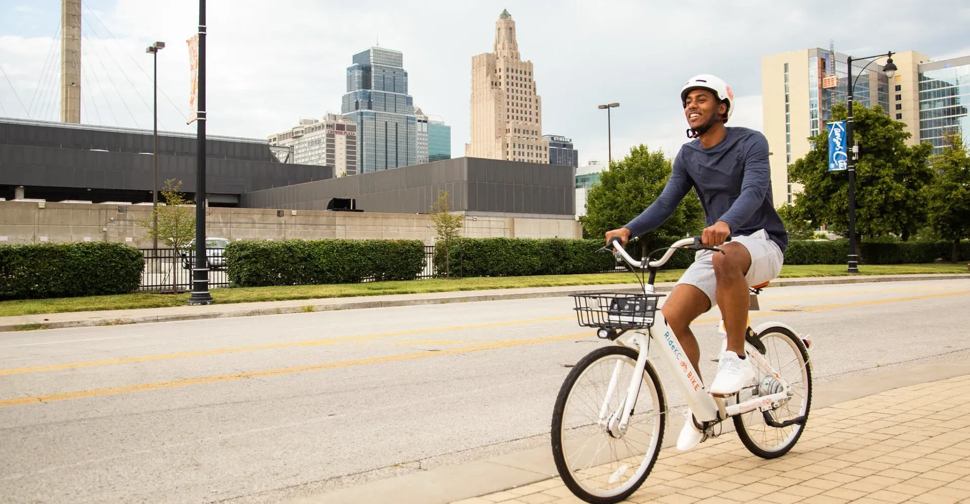 Person riding a white bicycle on a city street. Tall buildings are in the background.