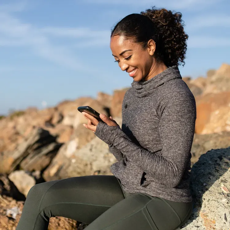 Woman in athletic wear looks at her phone while sitting outdoors on rocks.