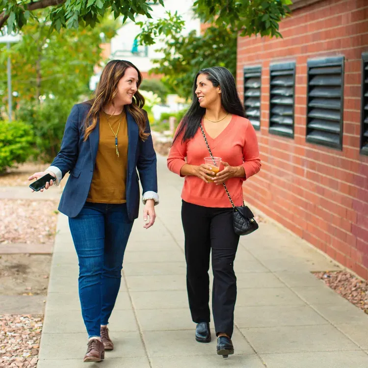 Two women walking and talking on a sidewalk, smiling. One holds a drink, the other a phone; brick building backdrop.