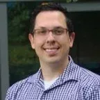 Man wearing glasses smiles, in a blue and white checkered shirt, outdoors.