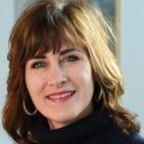 Woman with brown and blonde hair, smiling, wearing a dark turtleneck sweater, hoop earrings, indoors.