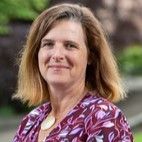 Woman with shoulder-length blonde hair wearing a patterned purple top, smiling, outdoors.
