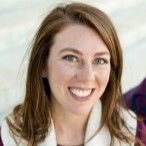 Woman smiling, brown hair, wearing a white vest, sitting outside.