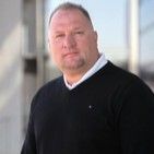 Man with short hair wearing a black sweater over a white collared shirt, outside a modern building.
