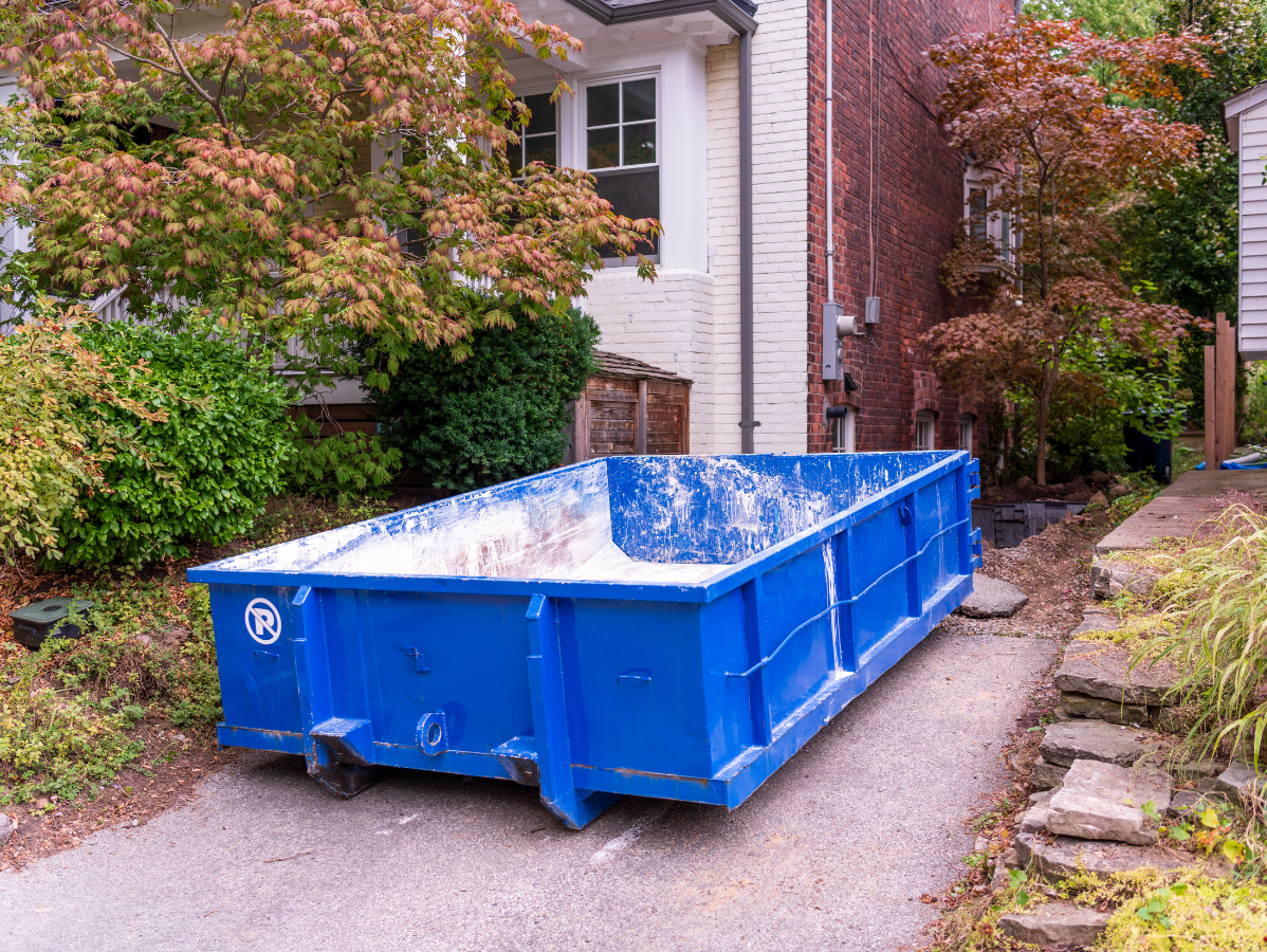 Blue dumpster in a driveway next to a brick building and a house with landscaping.