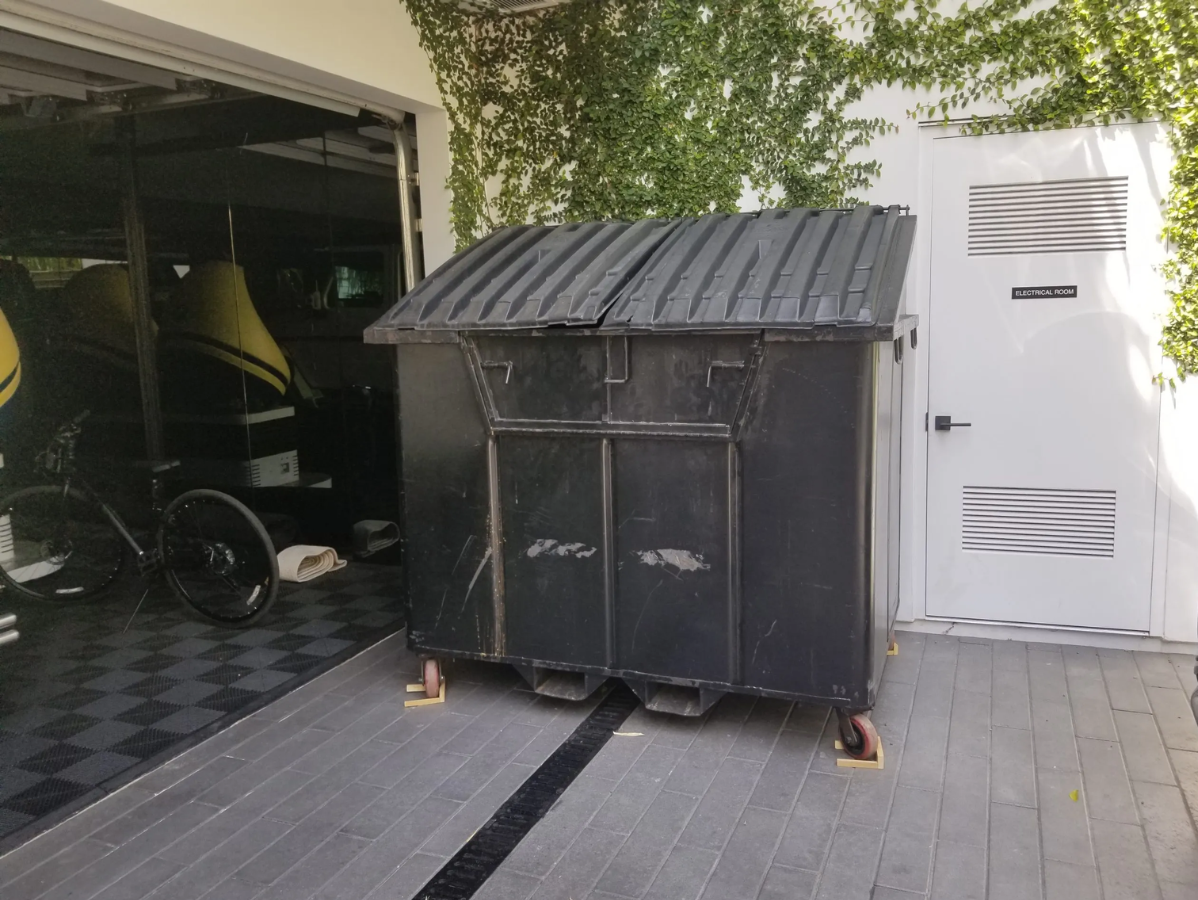 Black trash dumpster on a wooden deck next to a white door, next to a garage with a yellow car.