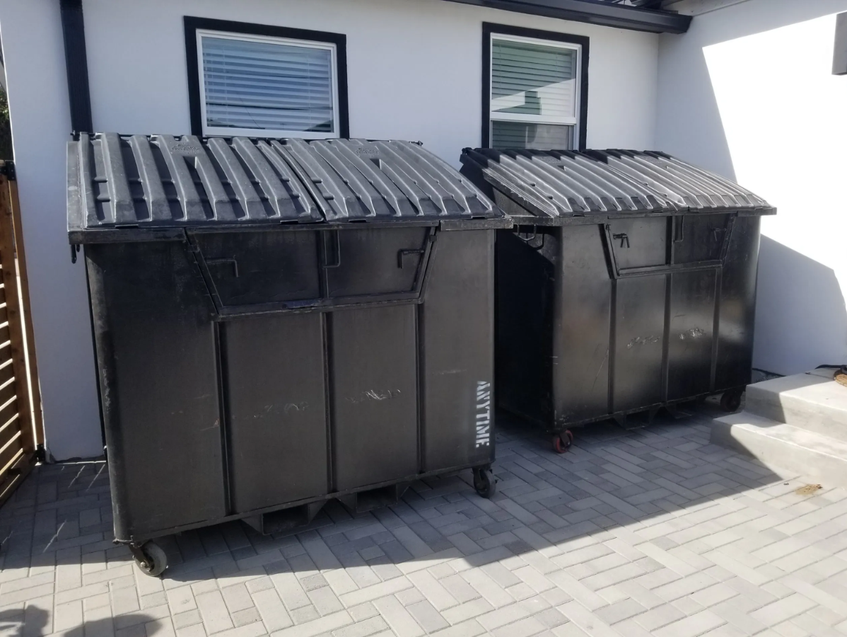 Two black dumpsters on a paved area, near a white building with windows.