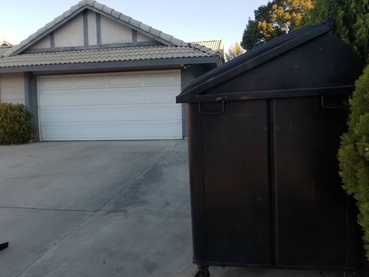 Black dumpster sits next to a home's garage; a tree is on the right.