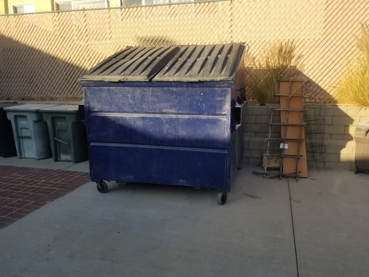 Blue dumpster with closed lid on concrete, near trash bins and a chain-link fence.