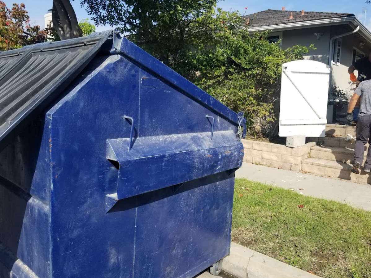Blue dumpster in front of a house, a person walks toward the door on a sunny day.