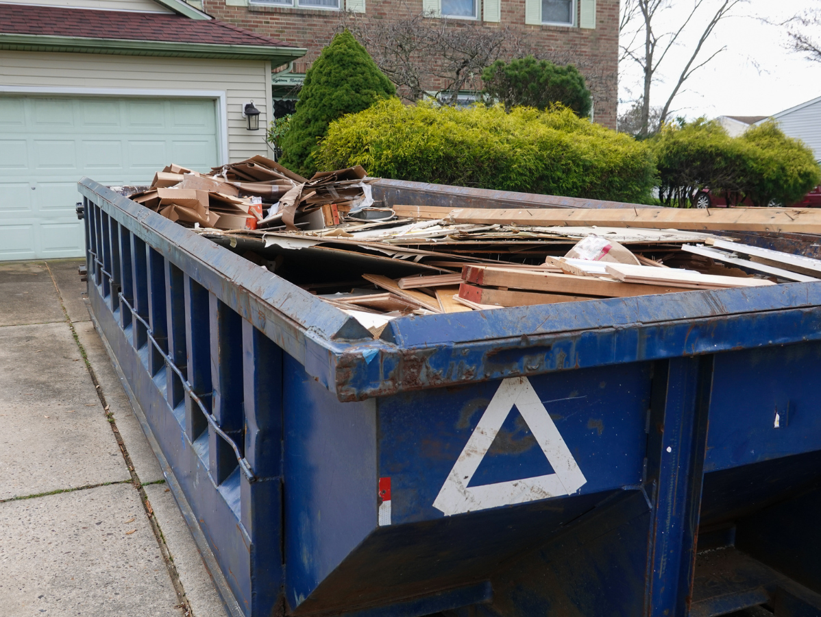 Blue dumpster filled with construction debris, parked on a driveway in front of a house.