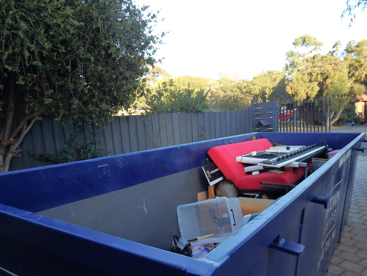 Blue dumpster overflowing with various trash; a red sofa cushion is visible.
