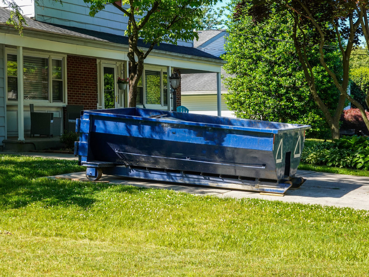 A blue dumpster rests on a concrete driveway in front of a house, surrounded by grass and trees.