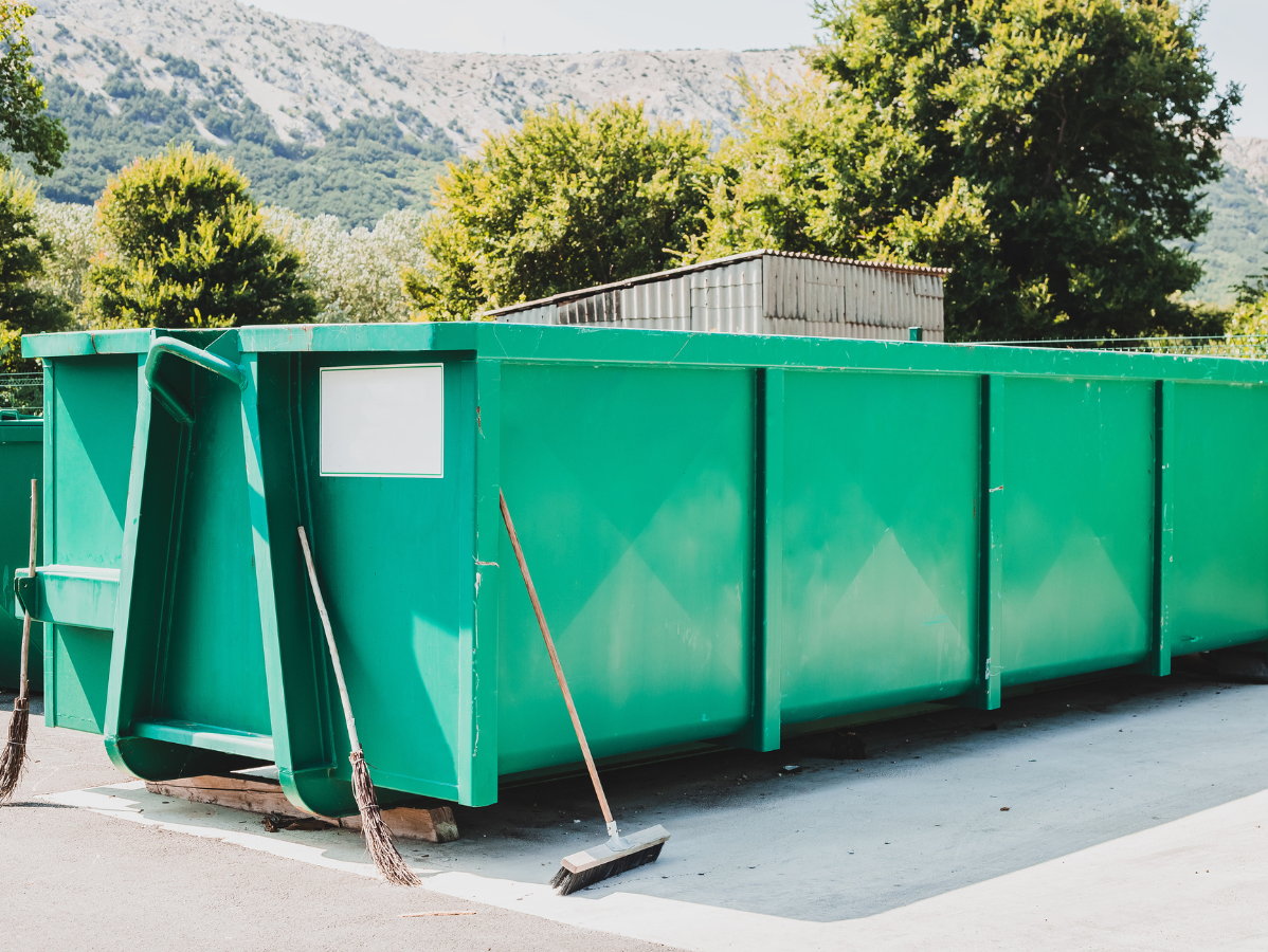 Green dumpster outdoors with brooms leaning against it, trees, and mountains in the background.