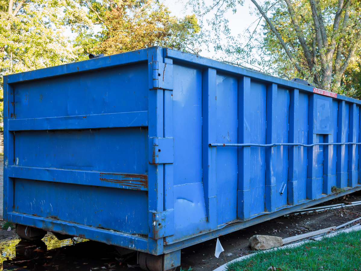 Blue dumpster on a driveway, set against a backdrop of trees and a green lawn.