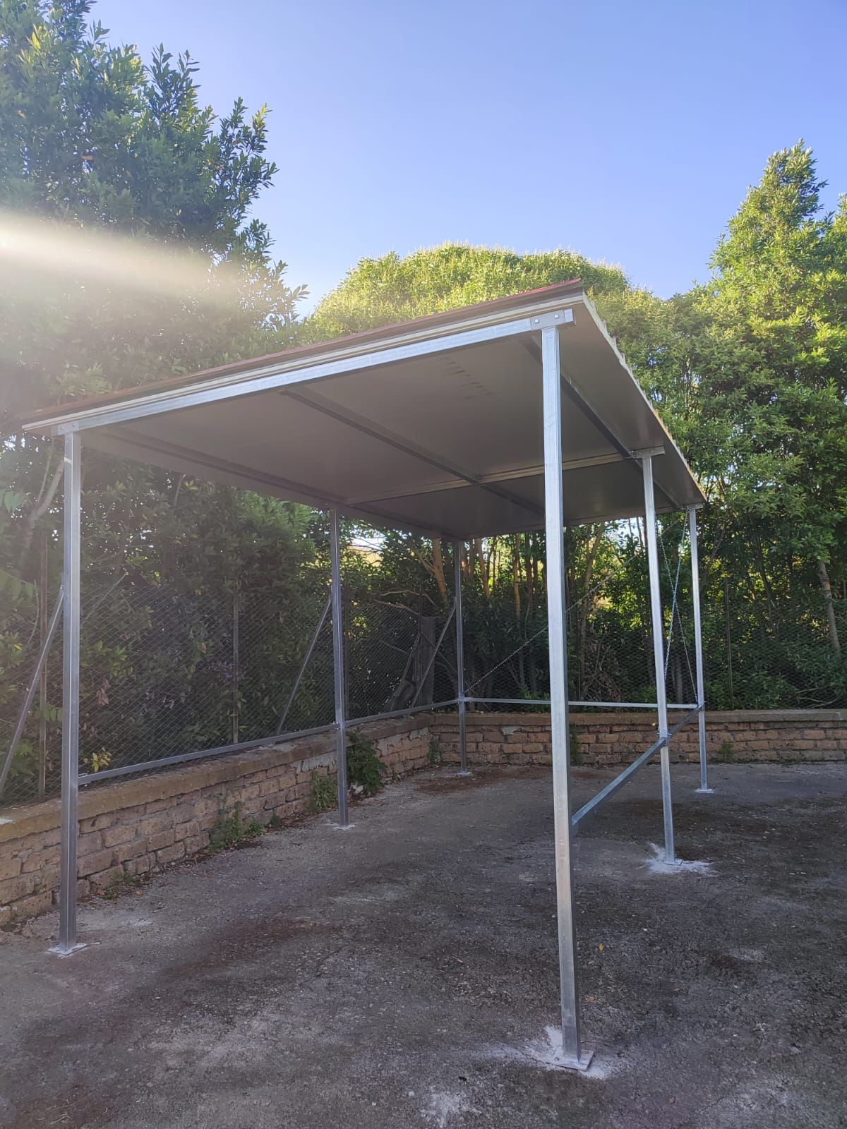 Struttura in metallo per posto auto coperto con tetto color marrone chiaro, base in cemento e alberi circostanti.