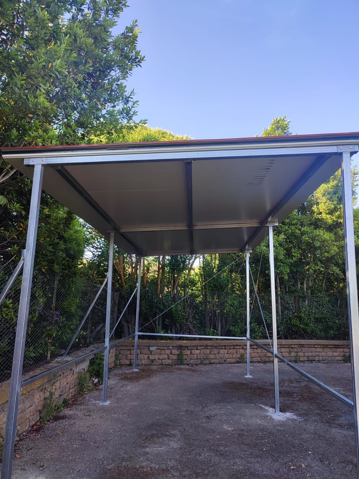 struttura in metallo e tetto in un ambiente esterno, probabilmente una fermata dell'autobus, con alberi e cielo limpido.