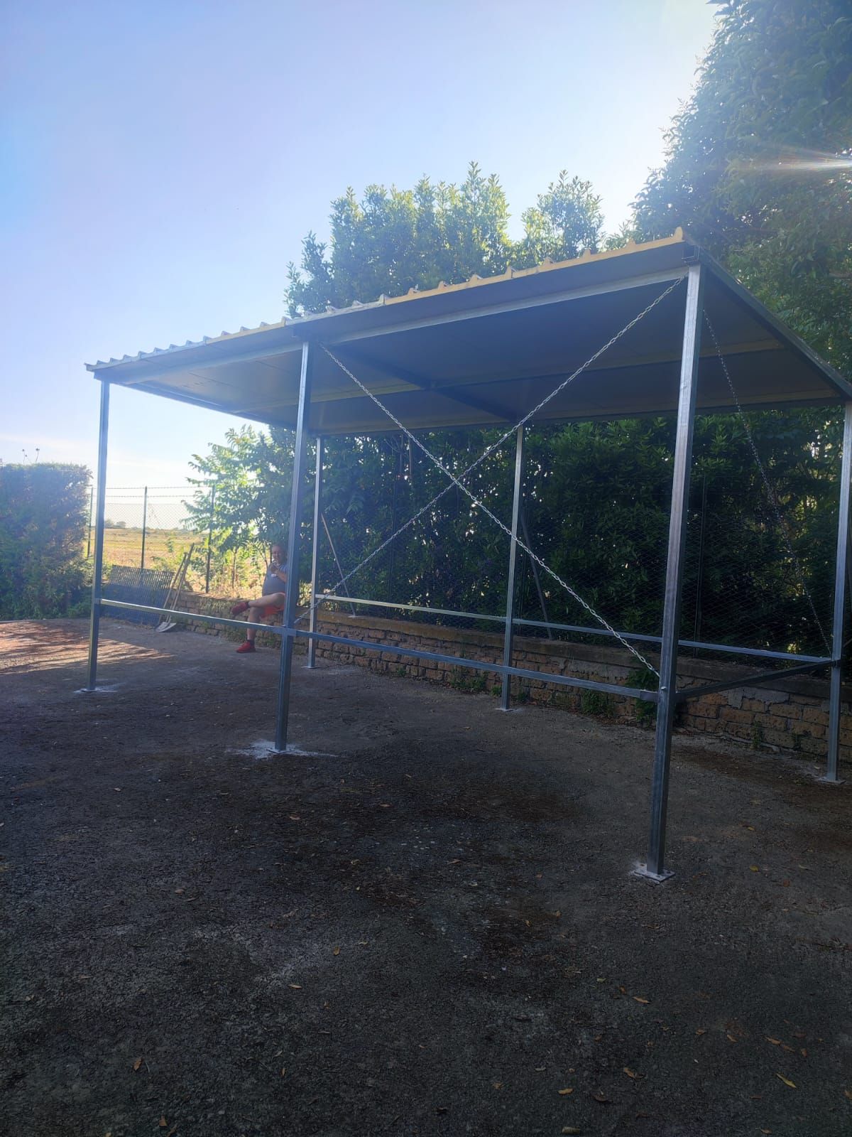 Rifugio in metallo con tetto grigio in una zona ghiaiosa, alberi sullo sfondo, giornata di sole.