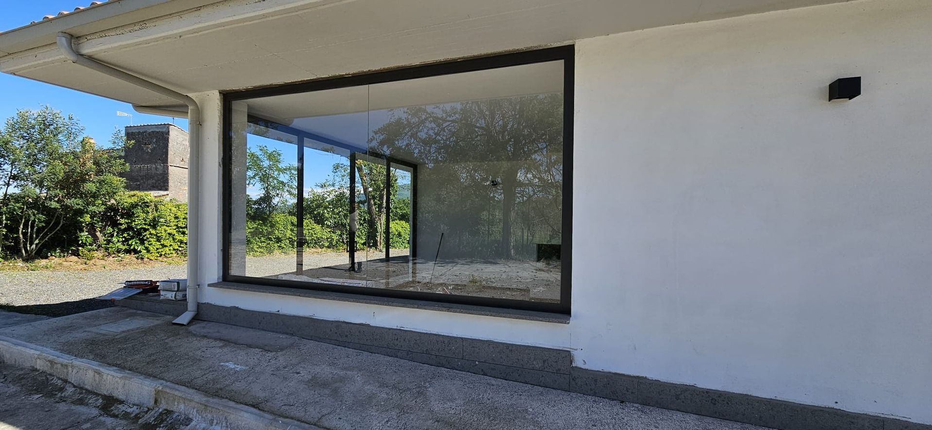 Esterno di un edificio moderno con grandi vetrate che riflettono gli alberi e un muro bianco con una lampada nera.